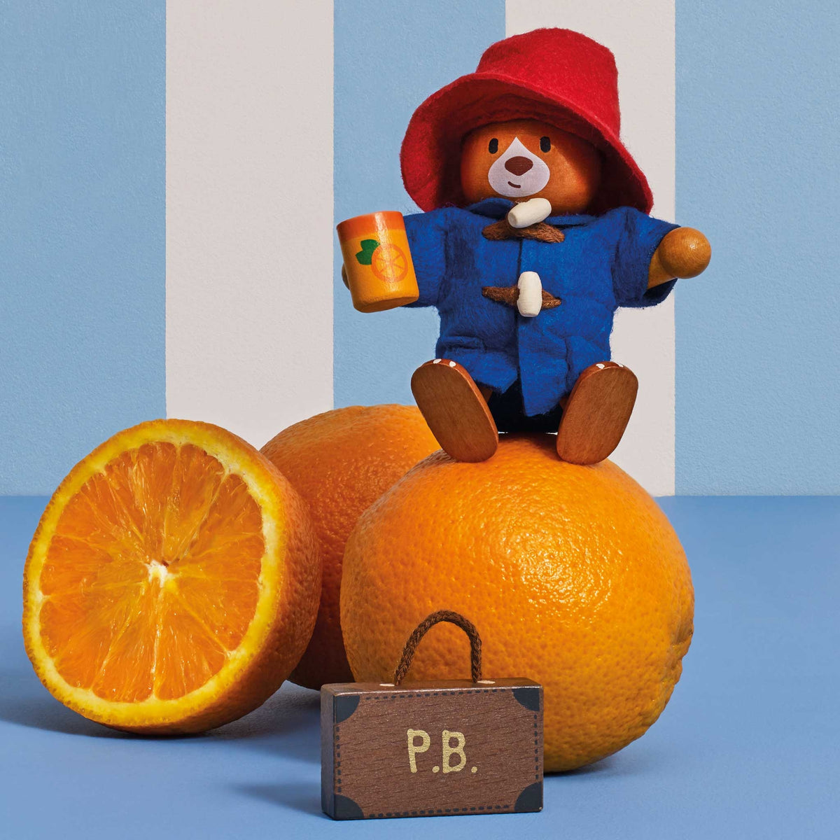 A Paddington Bear wooden doll wearing its signature red hat and blue duffel coat, sitting on top of a whole orange and holding an orange mug. A cut orange half and a small brown suitcase labeled 'P.B'