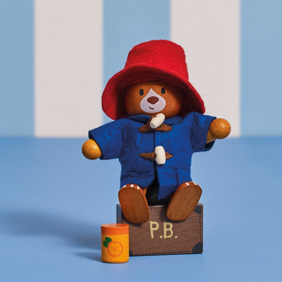 A wooden Paddington Bear doll wearing a red hat and blue coat, seated on a small brown box labeled P.B.