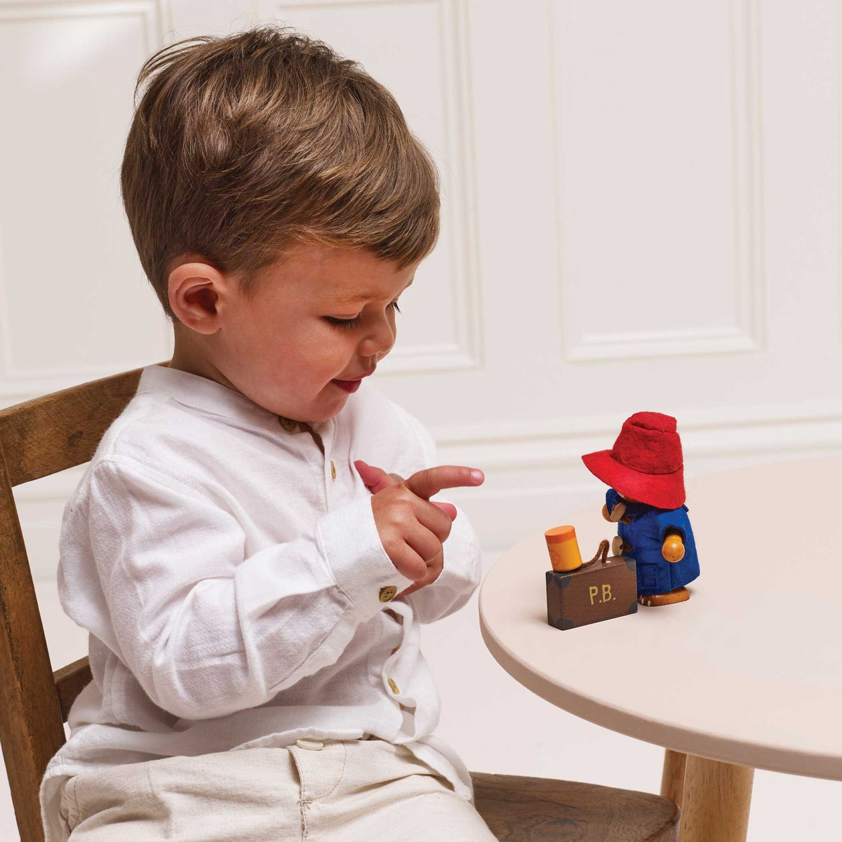 A toddler in a white long-sleeve shirt and beige trousers sits at a small round table, pointing toward a small Paddington toy figure with a red hat and brown suitcase.