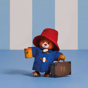 A Paddington Bear collectible figurine wearing a red hat and blue duffel coat, holding a jar of marmalade and a brown suitcase with 'P.B.