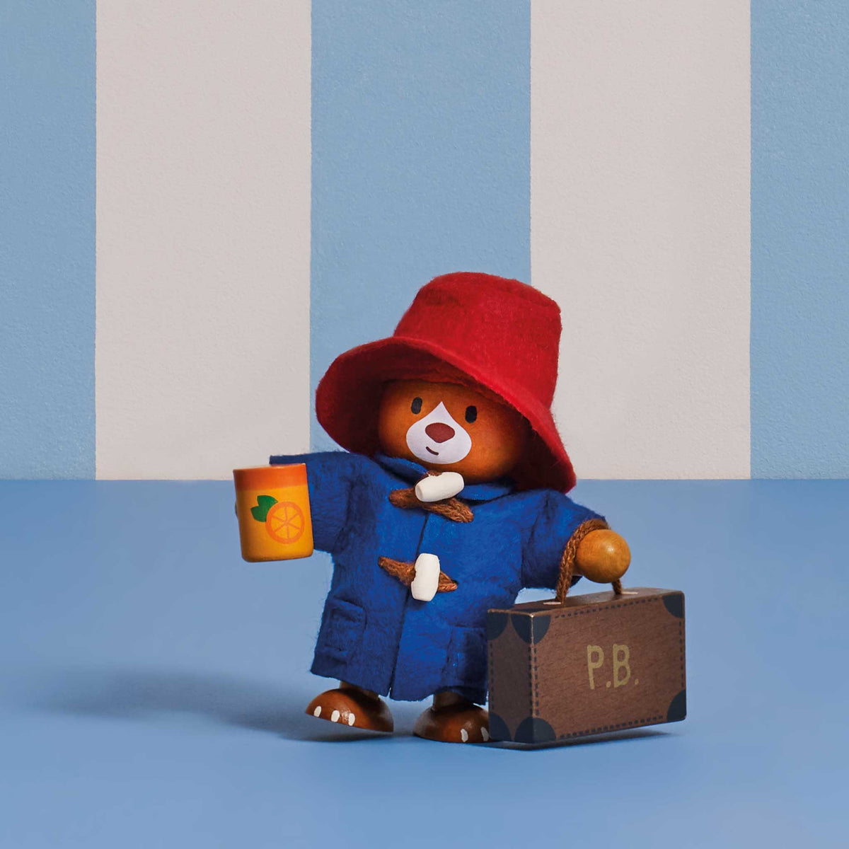 A Paddington Bear collectible figurine wearing a red hat and blue duffel coat, holding a jar of marmalade and a brown suitcase with 'P.B.