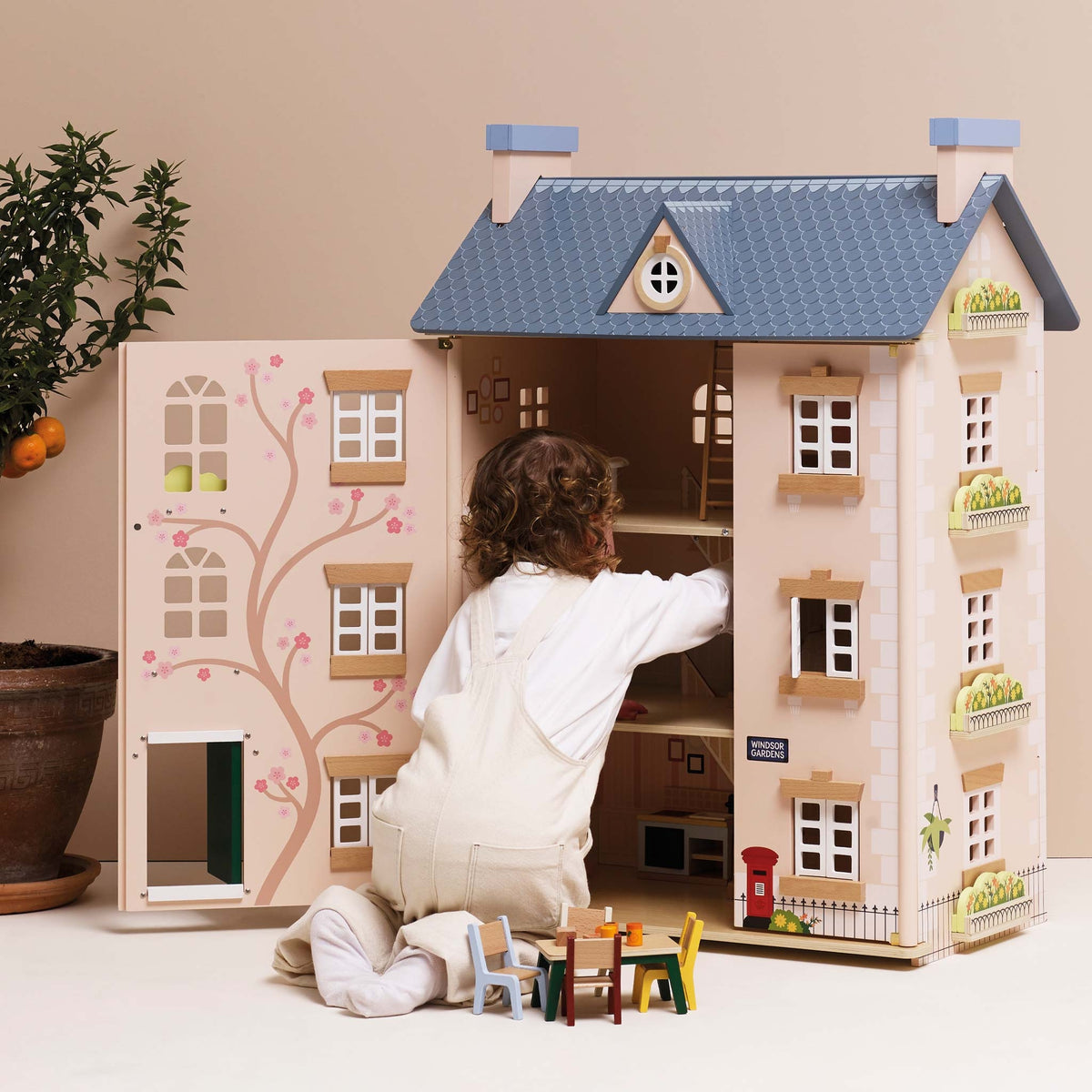 A child in cream overalls kneels beside a large wooden dollhouse with open side doors, reaching inside. The dollhouse has a blue-gray roof, cream walls, pink floral tree details, and small toy furniture and accessories.