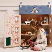 A young girl with curly hair sits inside a large wooden dollhouse playset, playing with a small table and toy figures. The dollhouse features a blue patterned roof, beige walls, and detailed interior rooms, set against a soft beige background.