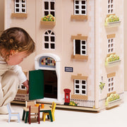 A young child in a cream outfit is playing with a large wooden dollhouse. The dollhouse is light beige with white brick-style detailing, multiple windows, a front door with a green panel, and small accessories including a red post box, black fence, and miniature furniture.