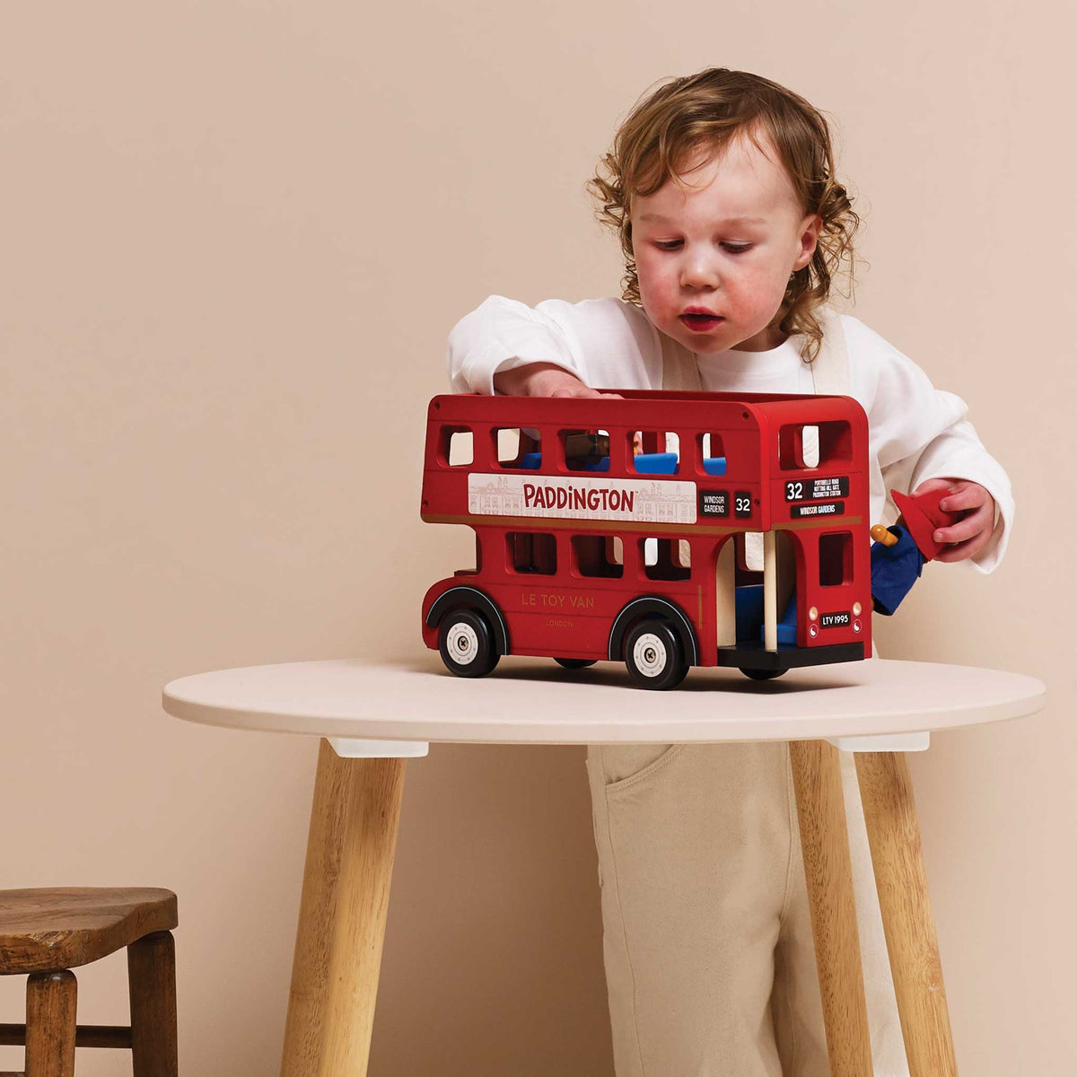 A toddler in a white long-sleeve top and beige pants sits behind a small round table, holding a red double-decker toy bus with 'PADDINGTON' written on the side. The scene is photographed in a warm, minimalist studio setting with a beige wall and light wood table.