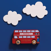 A red double-decker toy bus labeled 'LE TOY VAN' and 'PADDINGTON' is shown against a dark blue background with two white cloud cutouts above it.