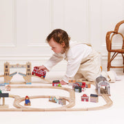 A toddler in a white outfit plays with a wooden toy train set on a white floor. The set includes curved wooden tracks, a red double-decker bus, bridge and station pieces, and small house models.