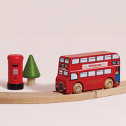 A wooden toy double-decker bus labeled 'LE TOY VAN' and 'PADDINGTON EXPRESS' sits on a curved wooden track, alongside a small red post box and green tree on a plain light beige background.