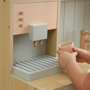 A close-up of a children's play kitchen or toy dispenser with a peach drawer, light wood frame, and pale gray drip tray, while a child's hands hold a matching small cup.