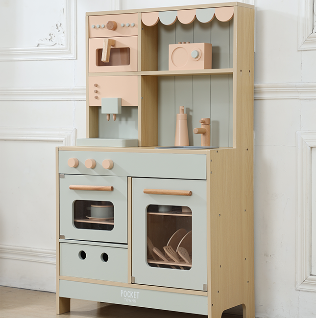 A children's wooden play kitchen set with a light natural wood frame and pale sage green panels, including oven doors, drawers, shelves, and small toy accessories, photographed against an off-white paneled wall.