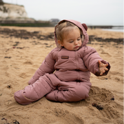 A toddler sitting on a sandy beach wearing a dusty rose puffer snowsuit with a matching hood.