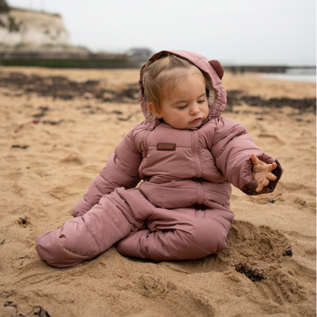 A toddler sitting on a sandy beach wearing a dusty rose puffer snowsuit with a matching hood.