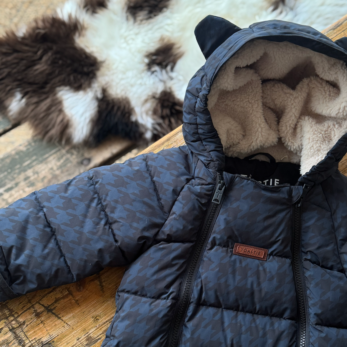 A navy blue puffer jacket with a subtle houndstooth pattern, featuring a plush cream fleece lining inside the hood with cat ears, laid flat on a rustic wooden surface.
