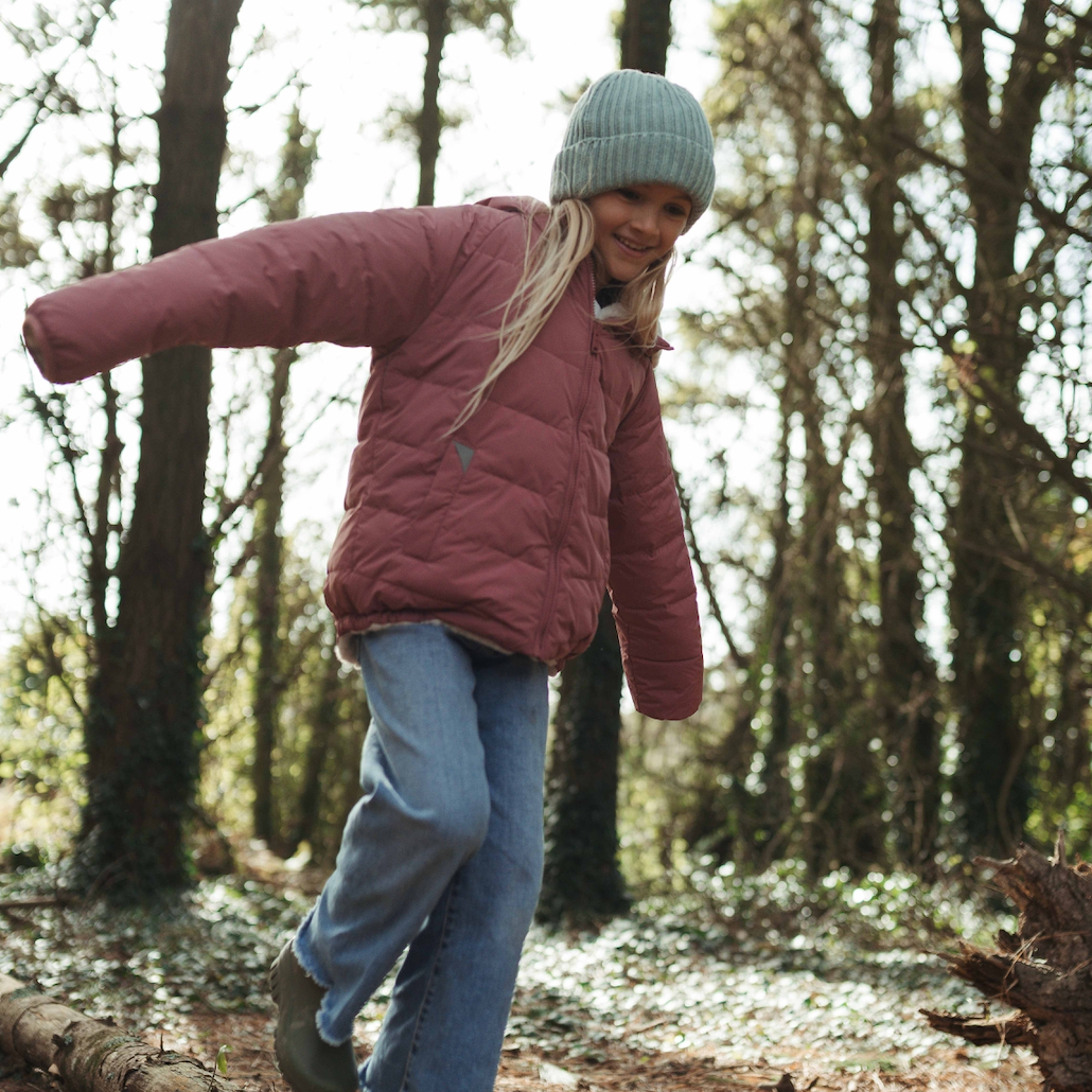 A girl wearing a dusty rose puffer jacket, blue jeans, a green knit beanie, and green boots is balancing on a fallen log in a forest.