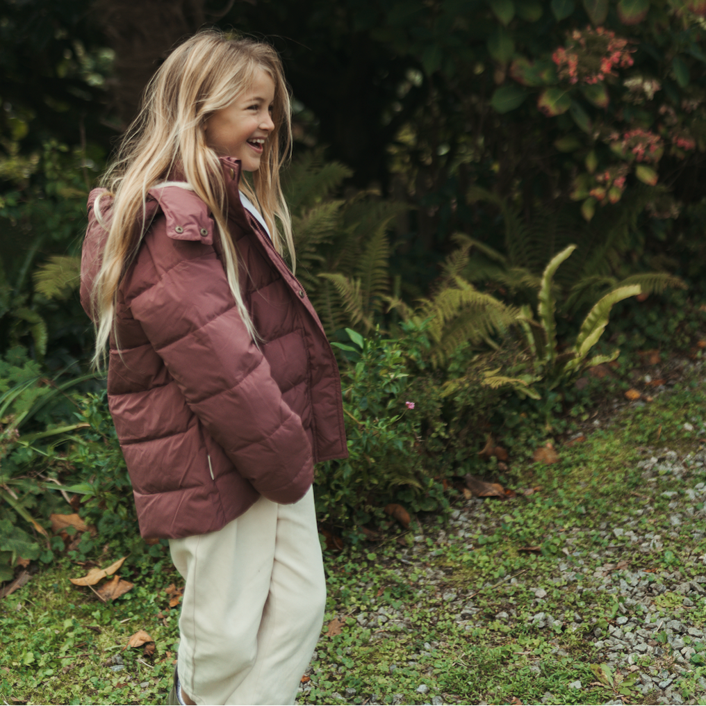 A young girl with long blonde hair stands outdoors in a garden wearing a dusty rose quilted puffer jacket and cream-colored pants, looking to the side with a smile.