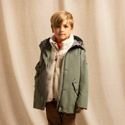 A young boy stands in a studio wearing an olive green hooded jacket over a cream zip-up fleece, brown pants, and white sneakers, posed against a draped beige fabric backdrop.