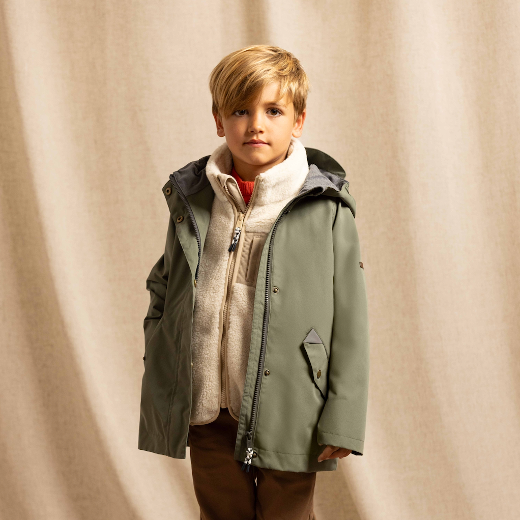 A young boy stands in a studio wearing an olive green hooded jacket over a cream zip-up fleece, brown pants, and white sneakers, posed against a draped beige fabric backdrop.