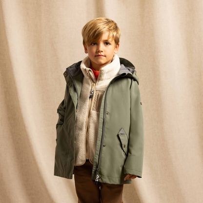 A young boy stands in a studio wearing an olive green hooded jacket over a cream zip-up fleece, brown pants, and white sneakers, posed against a draped beige fabric backdrop.