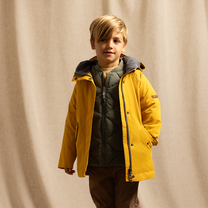 A young boy stands in front of a beige fabric backdrop wearing a bright yellow hooded jacket over a dark quilted puffer, brown pants, and black rain boots.