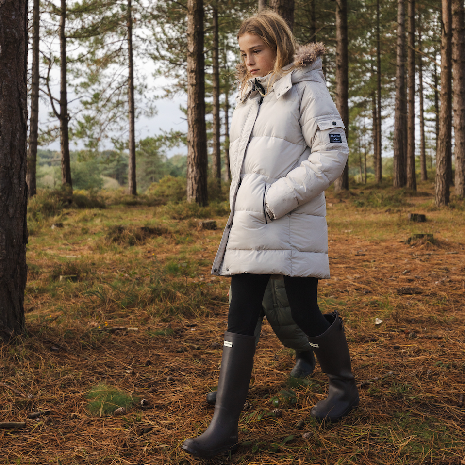 Girl walking through a forest wearing a long off white parka coat with the fur trim hood.