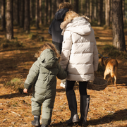 Three children wearing matching light olive green puffer snow suits walk hand in hand through a pine forest, with a dog nearby and another person in the background.