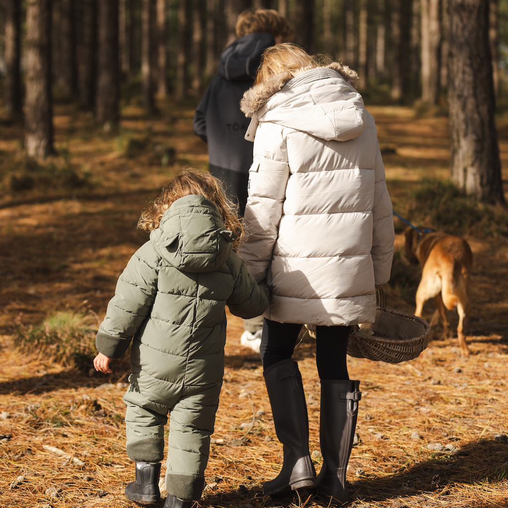 Three children wearing matching light olive green puffer snow suits walk hand in hand through a pine forest, with a dog nearby and another person in the background.