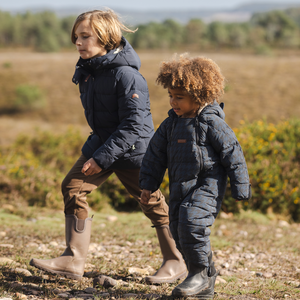 Two children walking outdoors in a field, wearing matching dark navy puffer jackets and brown pants. One child wears tall beige boots, and the other wears black boots.