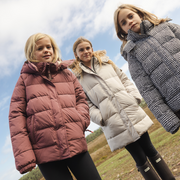 Three girls wearing different puffer jackets pose outdoors under a cloudy blue sky. The girl on the left wears a dusty rose puffer jacket, the middle girl wears a light beige puffer jacket, and the girl on the right wears a black and white houndstooth puffer jacket.
