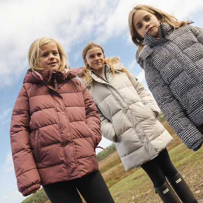 Three girls wearing different puffer jackets pose outdoors under a cloudy blue sky. The girl on the left wears a dusty rose puffer jacket, the middle girl wears a light beige puffer jacket, and the girl on the right wears a black and white houndstooth puffer jacket.