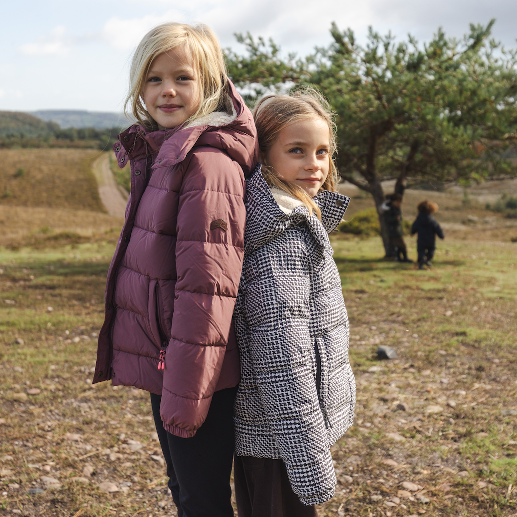 Two young girls standing outdoors in a grassy field under a cloudy sky, wearing quilted winter jackets. The girl on the left wears a plum-colored puffer jacket, and the girl on the right wears a black-and-white houndstooth patterned puffer jacket.