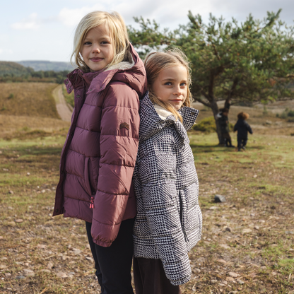 Two young girls standing outdoors in a grassy field under a cloudy sky, wearing quilted winter jackets. The girl on the left wears a plum-colored puffer jacket, and the girl on the right wears a black-and-white houndstooth patterned puffer jacket.