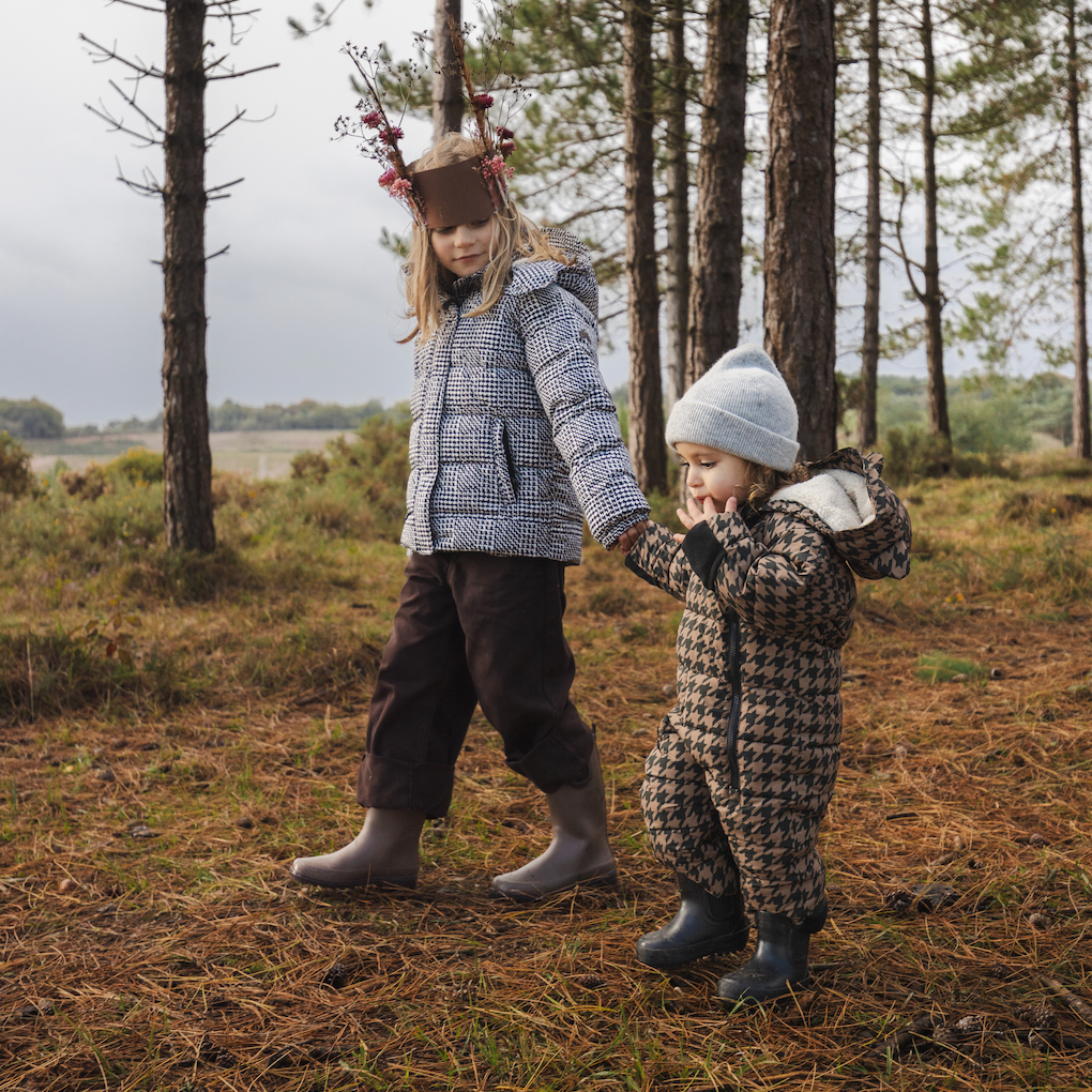 Two children walking hand in hand through a pine forest. The older child wears a grey quilted puffer jacket, dark brown trousers, and grey boots, with a reindeer antler headband.