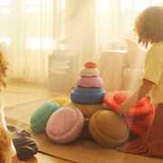 A child in a yellow shirt and rust skirt sits on a jute rug in a sunlit living room, playing with a stack of colourful Stapelstein Stepping Stones.