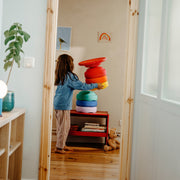A child stands in a doorway holding a stack of colourful stack of stapelstein stepping stones in a bright room with wood floors and toys.