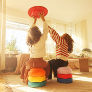 Two children sitting on colourful stapelstein stones in a sunlit living room, playing with a red wobble board. The children are seen from behind, wearing cosy knit sweaters, with warm golden sunlight streaming through the curtains.