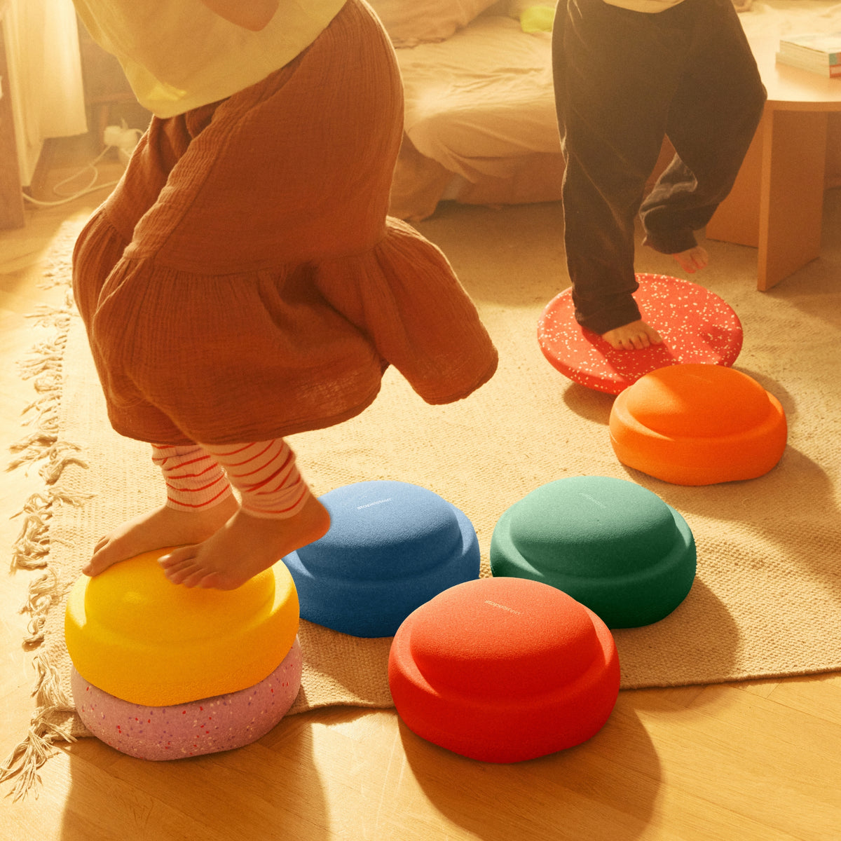 Children playing with a set of colourful stapelstein stones on a wooden floor in a sunlit room. The stools are stacked in different colours including yellow, pink, blue, green, orange, and red.