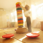 A child stands barefoot indoors next to a very tall stacked tower of brightly colored Stapelstein Stepping Stones.