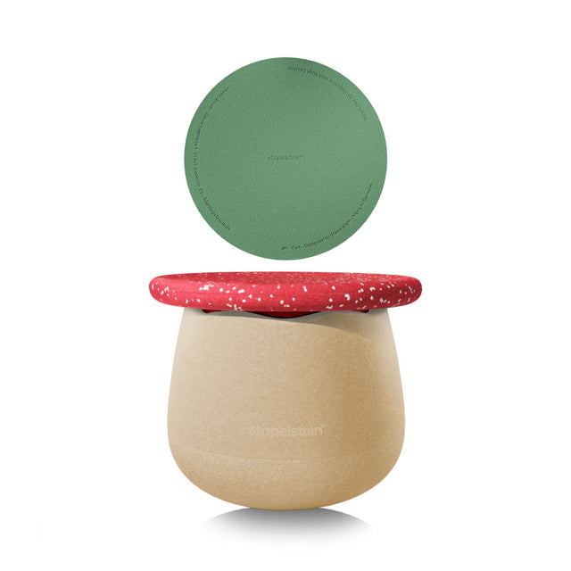 A Stapelstein Dynamic Set storage stool with a beige textured base, a red speckled wobble board top, and a green circular pad, shown on a white background.