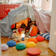 Two children sitting inside a fabric play tent on a wooden floor, with colourful stepping stones and a small blue lamp in a bright room.