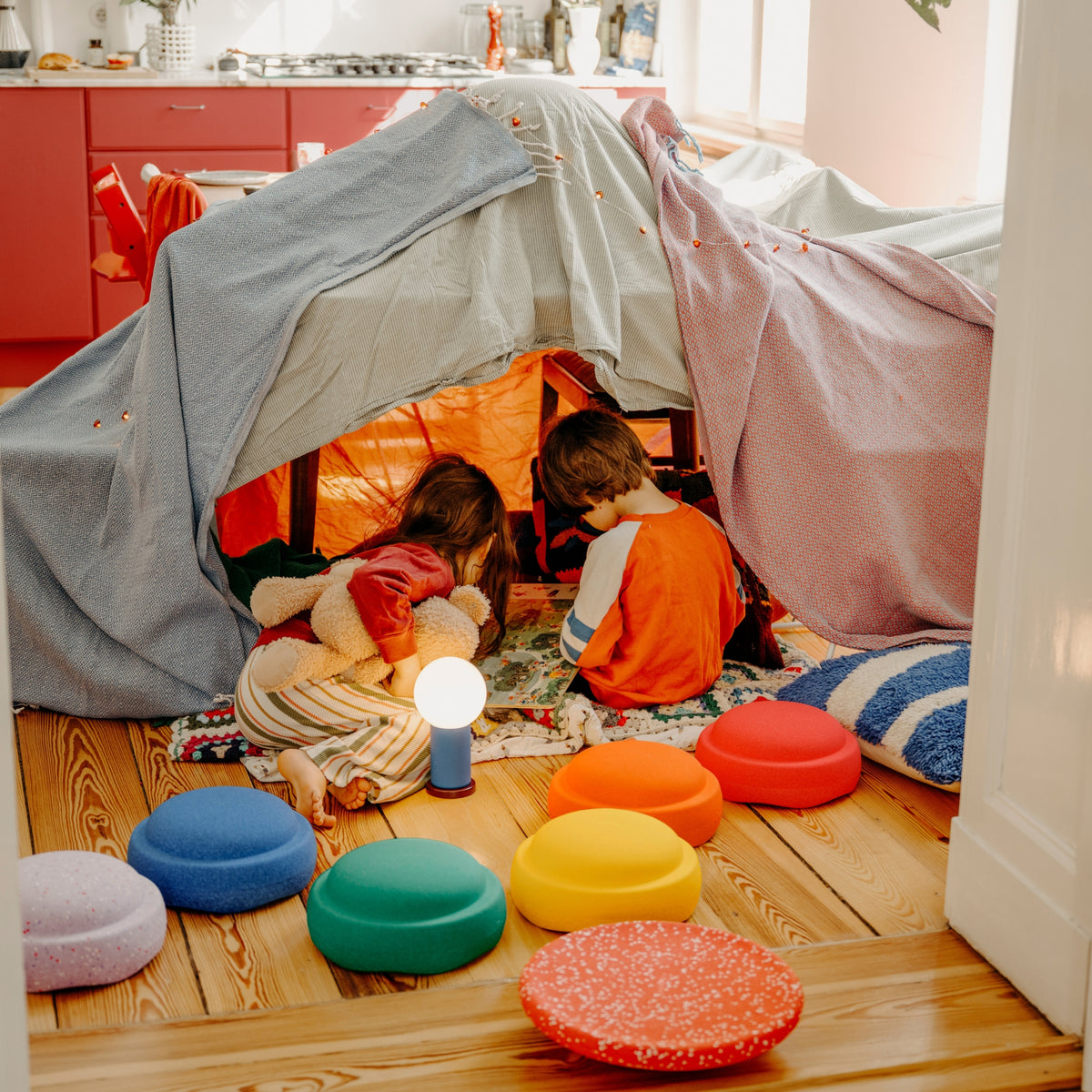Two children sitting inside a fabric play tent on a wooden floor, with colourful stepping stones and a small blue lamp in a bright room.