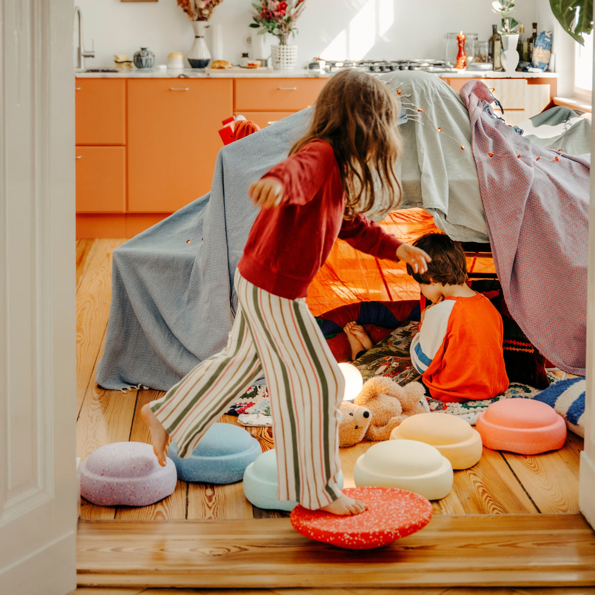 A child in a red top and striped trousers walks barefoot over round colourful stepping stones while another child sits inside a play tent made from blankets in a bright room with orange cabinets and wood floors.