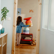 A child stands barefoot in a child's room, stacking colourful soft stepping stones. The room has light wood flooring and walls, with a small plant on a shelf to the left and a teddy bear on the floor.