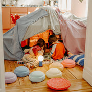 Two children play inside a cosy fabric play tent set up on a wooden floor, with colourful stepping stones arranged around it. 