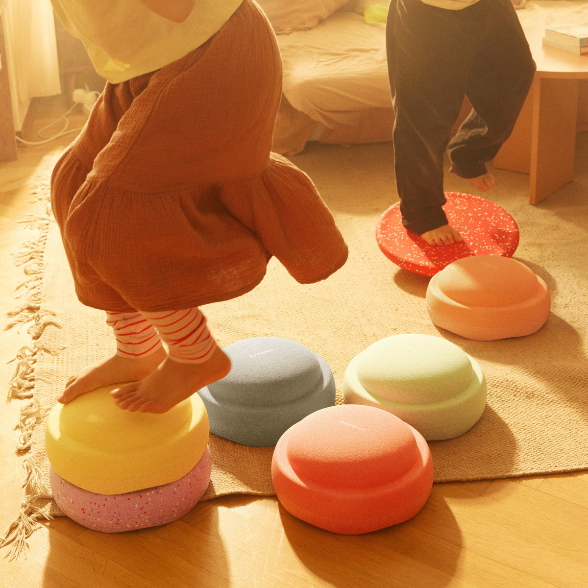 Two children stepping on round, pastel Stapelstein balance stepping stones arranged on a rug in a warm, sunlit room.
