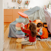 Two children playing in a cosy indoor play scene with a fabric tent and a stack of pastel stepping stone toys on a wooden floor.