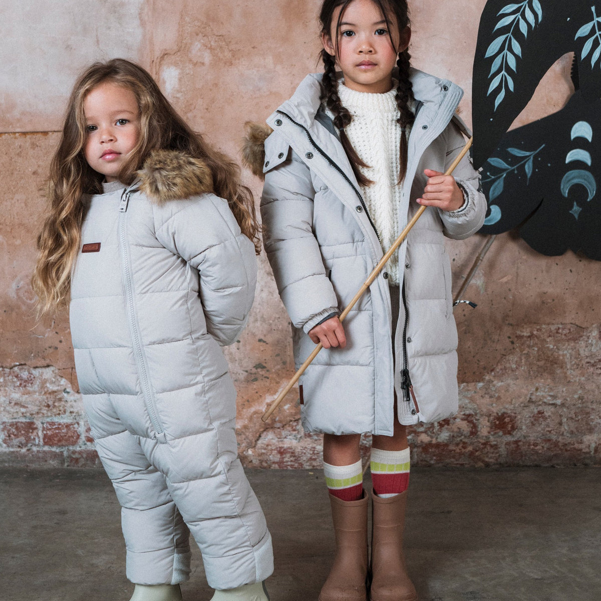 Two young girls modeling white puffer snowsuits in a rustic indoor setting with a textured plaster wall. The girl on the left wears a full-body snow suit with a faux-fur collar and white rubber boots.
