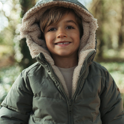 A smiling young boy wearing an olive green hooded puffer jacket with a beige sherpa fleece lining, photographed outdoors in a softly blurred natural setting.