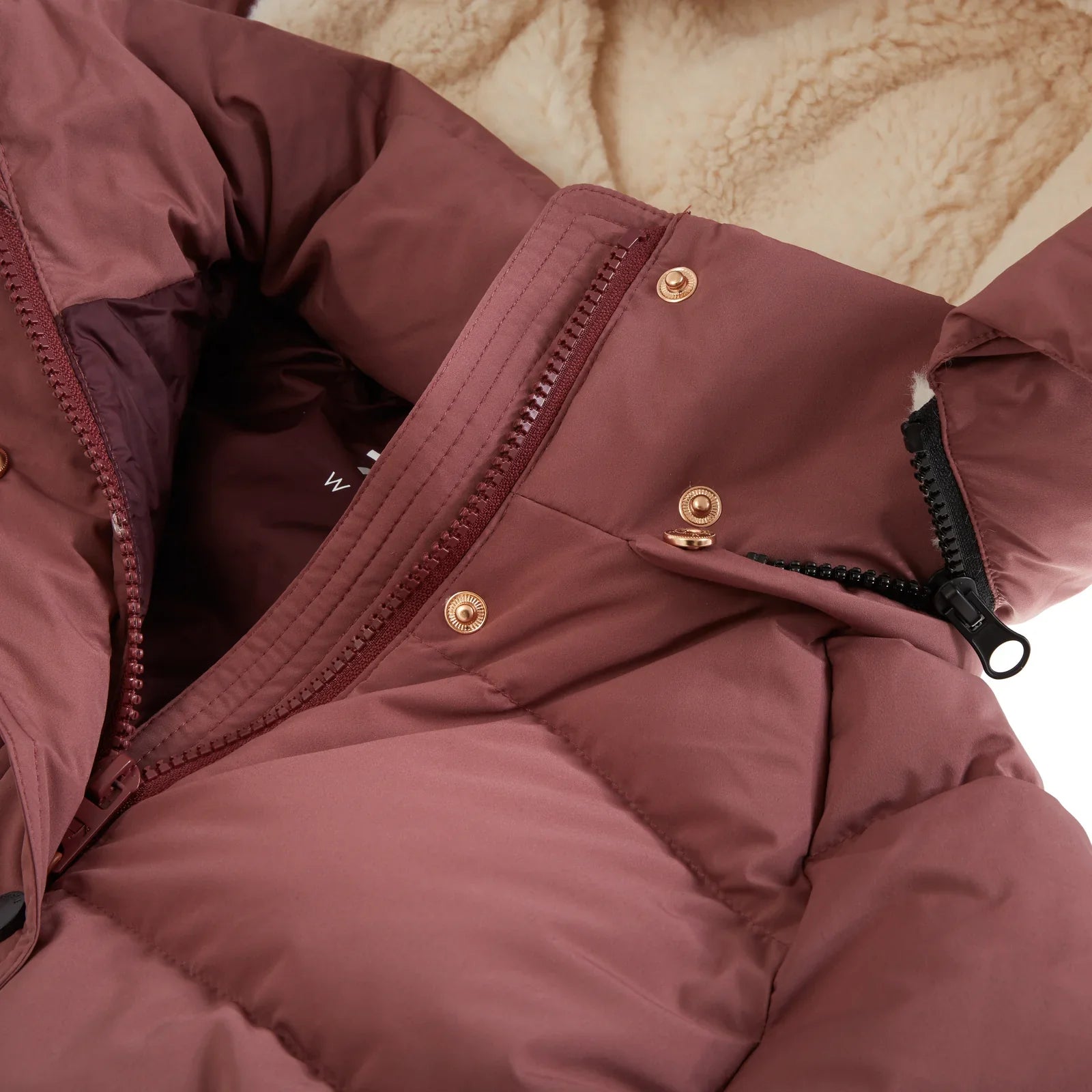 Close-up of a dusty rose puffer jacket showing the quilted fabric, gold snap buttons, a black zipper, and a beige sherpa lining.