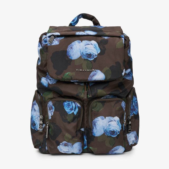 A Tiba + Marl dark brown backpack featuring a blue rose floral print over a subtle camouflage pattern, with multiple zippered compartments and a top handle, shown against a plain white background.