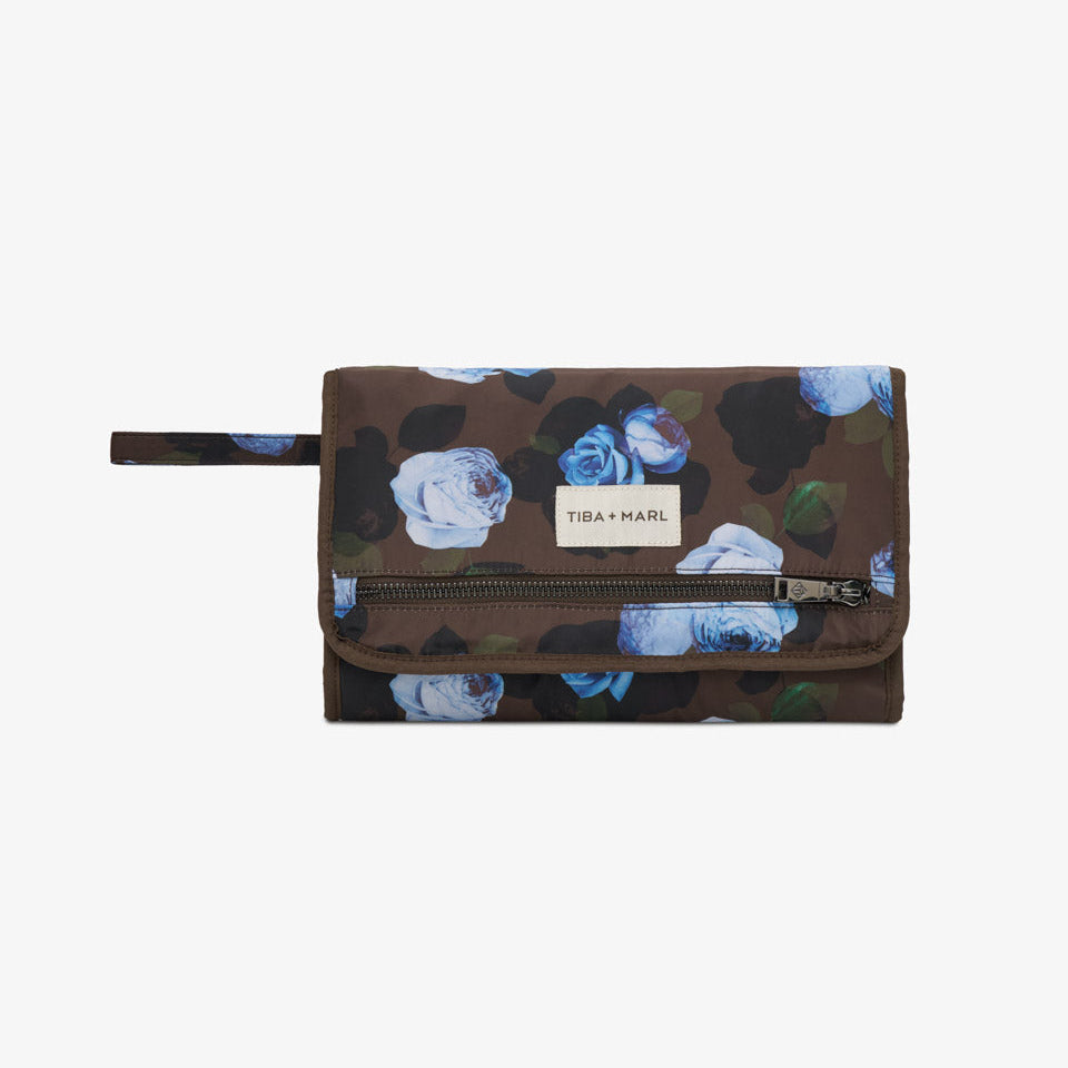 A Tiba + Harel zip-around wallet in a dark brown camouflage pattern with blue and white floral prints, featuring a white brand label on the front flap.